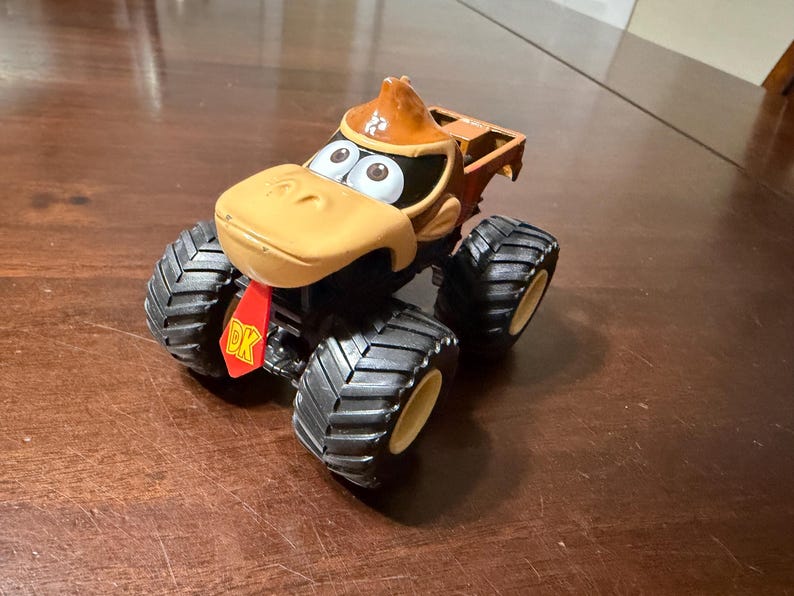 May include: A toy monster truck featuring a Donkey Kong character design. The truck has large black tyres with yellow rims, a tan face, and a brown hat. The red DK logo is visible on the front.