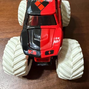 May include: A red and black toy monster truck with large white tires. The truck has the words "Harley Quinn" printed on the side. The truck is a toy vehicle.