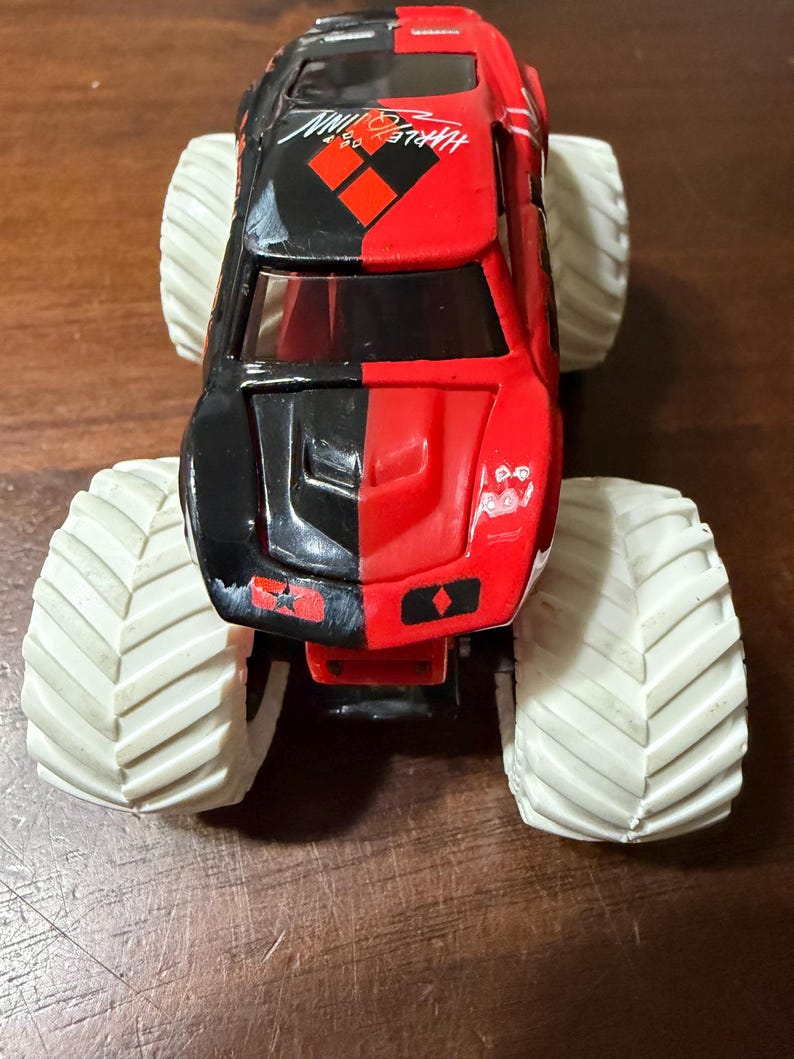 May include: A red and black toy monster truck with large white tires. The truck has the words "Harley Quinn" printed on the side. The truck is a toy vehicle.