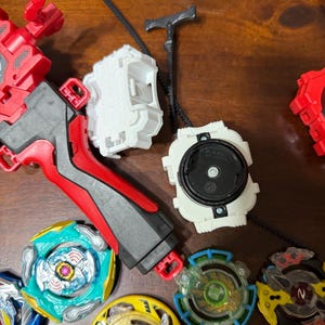 May include: A collection of colorful spinning top toys and launchers. The tops feature various designs with blue, green, yellow, and red accents. Launchers are red, white, and black. The toys are designed for competitive play.