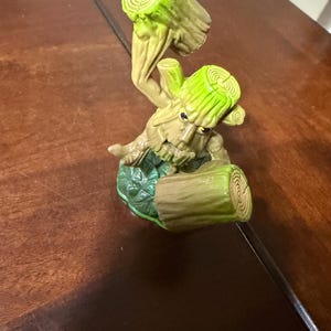 May include: A whimsical figurine of a tree-like creature with a light brown body and bright green accents. The figure has a face and is holding two log-shaped pieces. The base is green with leaf details. The overall design is cartoonish.