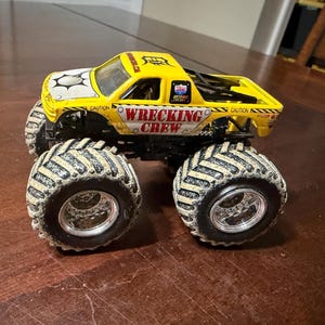Puede incluir: Un cami&oacute;n monstruo de juguete amarillo con las palabras "WRECKING CREW" en el lateral. El cami&oacute;n tiene neum&aacute;ticos negros grandes con un dibujo beige. La cabina tiene un dise&ntilde;o blanco y negro en el cap&oacute;.