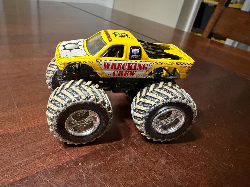 Puede incluir: Un cami&oacute;n monstruo de juguete amarillo con las palabras "WRECKING CREW" en el lateral. El cami&oacute;n tiene neum&aacute;ticos negros grandes con un dibujo beige. La cabina tiene un dise&ntilde;o blanco y negro en el cap&oacute;.