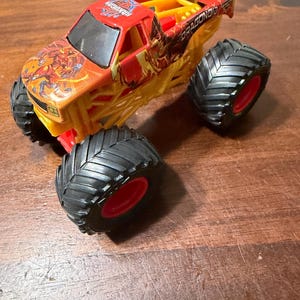 K&ouml;nnte beinhalten: Ein Spielzeug-Monstertruck in Rot, Gelb und Orange. Der Truck hat gro&szlig;e schwarze Reifen mit roten Felgen. Auf der Kabine steht das Wort "BakuGan" und auf der Seite "DRAGONOID". Der Truck steht auf einer Holzoberfl&auml;che.
