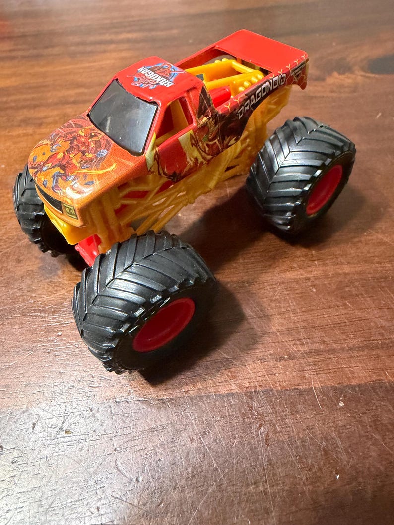 K&ouml;nnte beinhalten: Ein Spielzeug-Monstertruck in Rot, Gelb und Orange. Der Truck hat gro&szlig;e schwarze Reifen mit roten Felgen. Auf der Kabine steht das Wort "BakuGan" und auf der Seite "DRAGONOID". Der Truck steht auf einer Holzoberfl&auml;che.