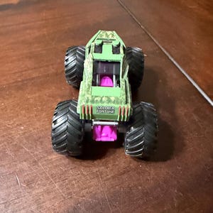 May include: A green and black toy monster truck with large black tires. The truck has a camouflage pattern on the top and the text "SOLDIER FORTUNE" on the back. The truck has a pink interior.