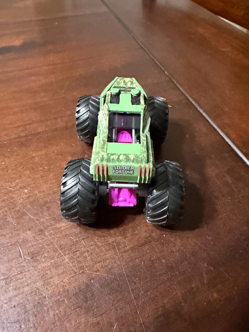 May include: A green and black toy monster truck with large black tires. The truck has a camouflage pattern on the top and the text "SOLDIER FORTUNE" on the back. The truck has a pink interior.