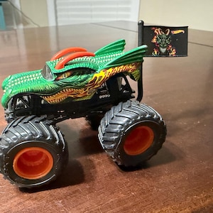May include: A green and orange monster truck toy with a dragon design. The truck has large black tires with orange rims and a small flag with a dragon graphic. The body of the truck is green with orange and yellow accents.