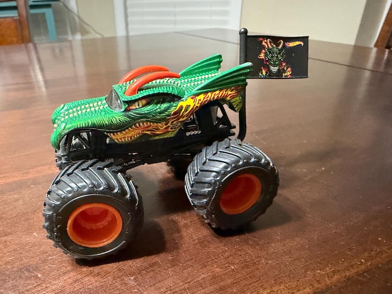 May include: A green and orange monster truck toy with a dragon design. The truck has large black tires with orange rims and a small flag with a dragon graphic. The body of the truck is green with orange and yellow accents.
