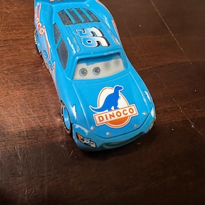 May include: A light blue toy race car with the word "DINOCO" on the spoiler and front. The car has the number "95" on the roof and side, and a dinosaur logo on the front. The car is a toy.