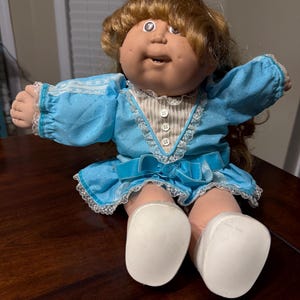 May include: A vintage doll with a soft, vinyl face and light brown hair. The doll wears a blue dress with white lace trim and a matching bow. The doll's shoes are white.