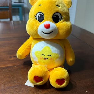 Puede incluir: Un oso de peluche amarillo con una cara sonriente, una nariz en forma de corazón rojo y una estrella en el vientre. El oso tiene un mechón de pelo amarillo y detalles en forma de corazón rojo en los pies. Un juguete tierno.