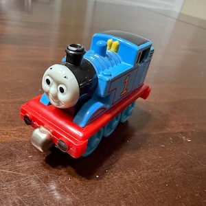 May include: A blue, red, and black toy train with the number 1 on the side. The train has a smiling face and is a classic children's toy.