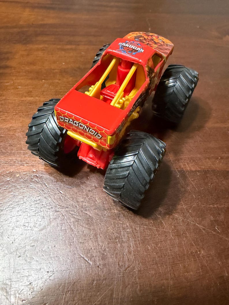 K&ouml;nnte beinhalten: Ein roter und gelber Spielzeug-Monstertruck mit gro&szlig;en schwarzen Reifen. Der Truck tr&auml;gt die Aufschrift "DRAGONOID" an der Seite und "BAKUGAN" oben.