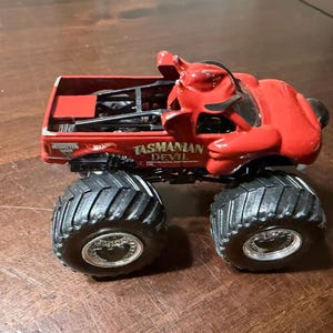 May include: A red toy monster truck with large black tires. The truck features a devil figure on top and the words "Tasmanian Devil" on the side. The truck is a collectible toy.