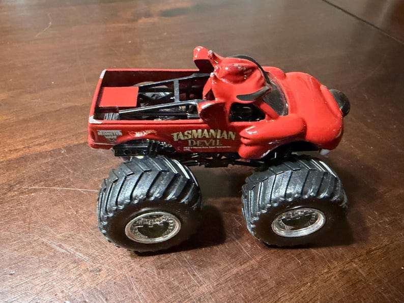May include: A red toy monster truck with large black tires. The truck features a devil figure on top and the words "Tasmanian Devil" on the side. The truck is a collectible toy.