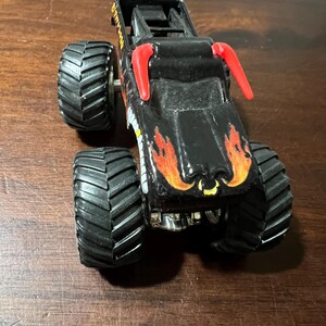 Op de afbeelding: Zwart monstertruckspeelgoed met grote, zwarte, getextureerde banden. De truck heeft oranje en gele vlammen op de motorkap en rode accenten. Het speelgoed is ontworpen met een gedetailleerd chassis en een rolkooi.
