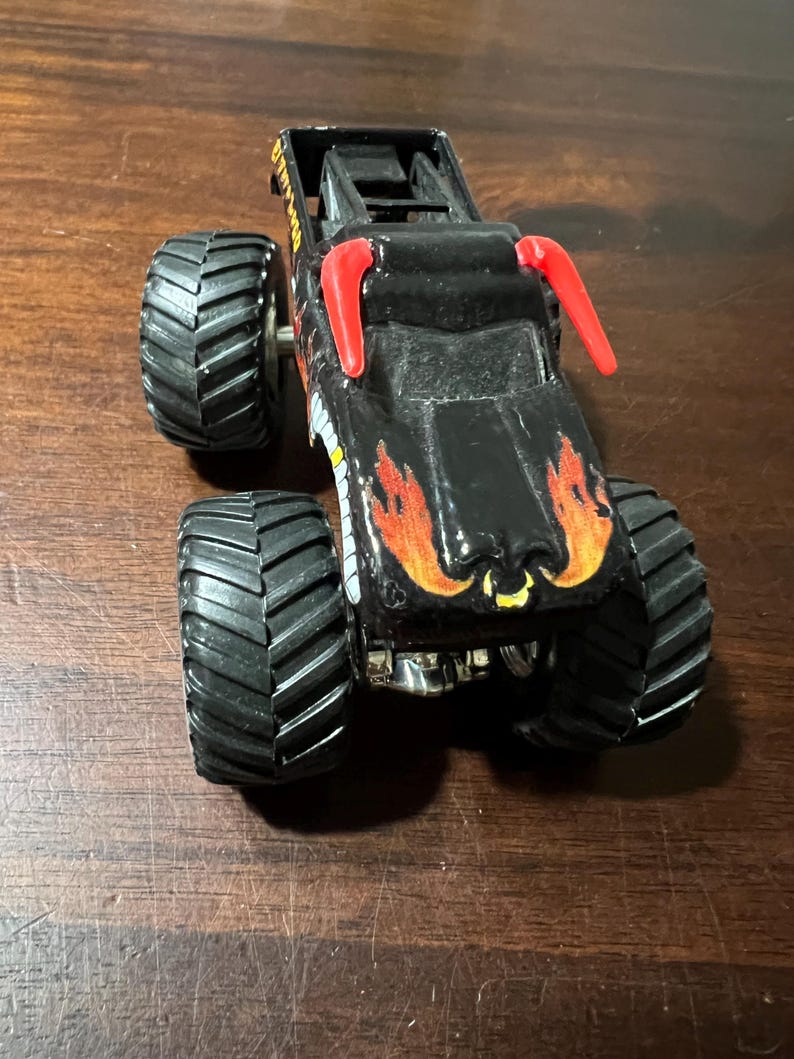 Op de afbeelding: Zwart monstertruckspeelgoed met grote, zwarte, getextureerde banden. De truck heeft oranje en gele vlammen op de motorkap en rode accenten. Het speelgoed is ontworpen met een gedetailleerd chassis en een rolkooi.