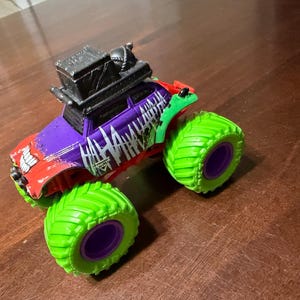May include: A toy monster truck with a red, purple, and green color scheme. The truck features large green tires with purple rims and the word "HA HA HA" in white. A black box is mounted on the roof.
