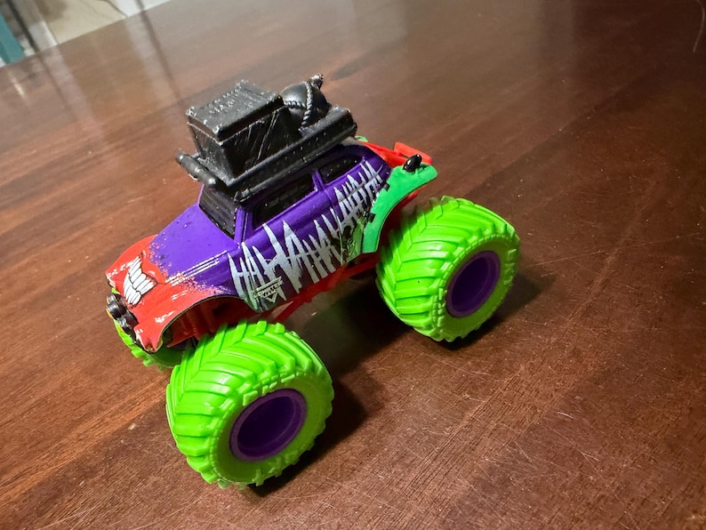 May include: A toy monster truck with a red, purple, and green color scheme. The truck features large green tires with purple rims and the word "HA HA HA" in white. A black box is mounted on the roof.
