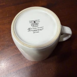May include: White ceramic mug with a curved handle. The bottom of the mug features text including "Designers Collection," "AMERICAN GREETINGS," and "ZIGGY." The mug is made of stoneware and is on a wooden surface.