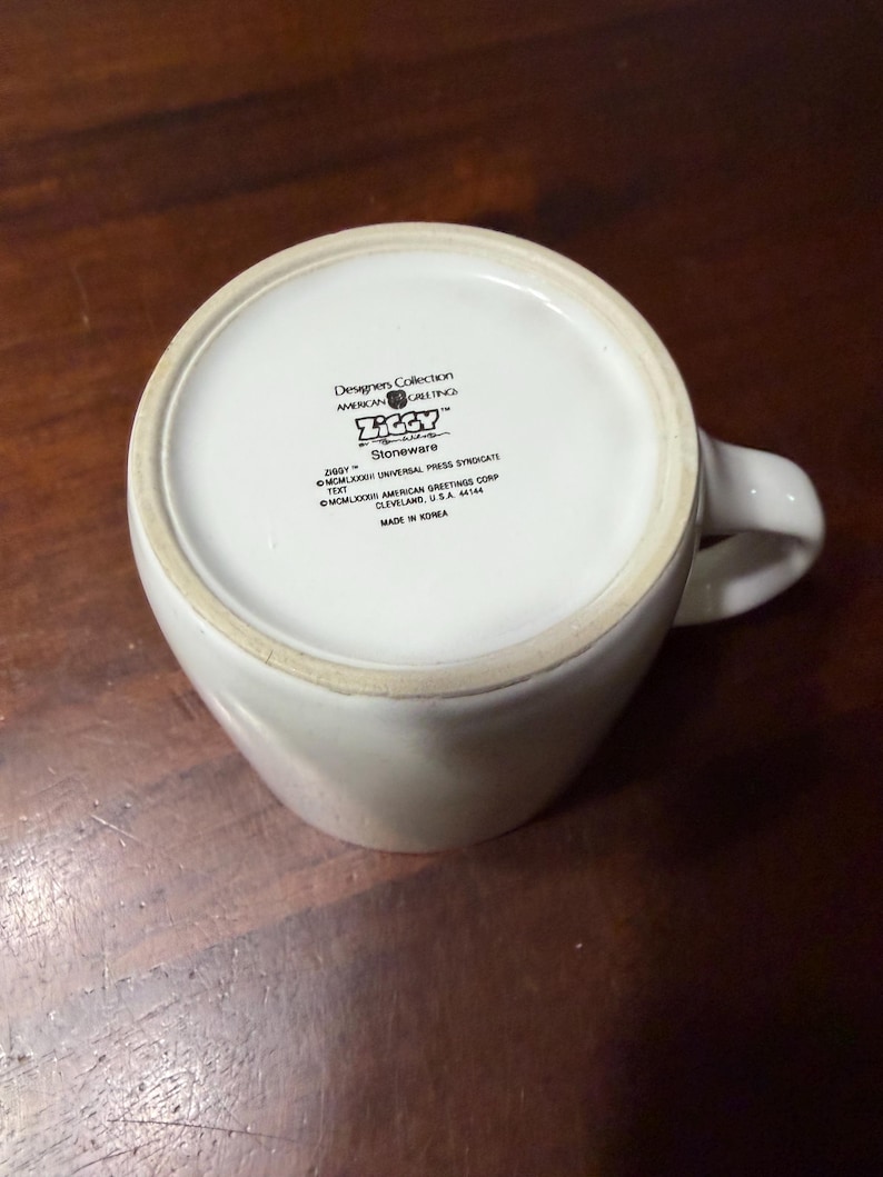May include: White ceramic mug with a curved handle. The bottom of the mug features text including "Designers Collection," "AMERICAN GREETINGS," and "ZIGGY." The mug is made of stoneware and is on a wooden surface.