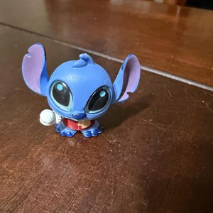 Pu&ograve; includere: Statuetta blu del personaggio Stitch del film d'animazione Lilo & Stitch. La statuetta ha grandi occhi neri, grandi orecchie e una piccola palla bianca in mano. Indossa un colletto rosso.