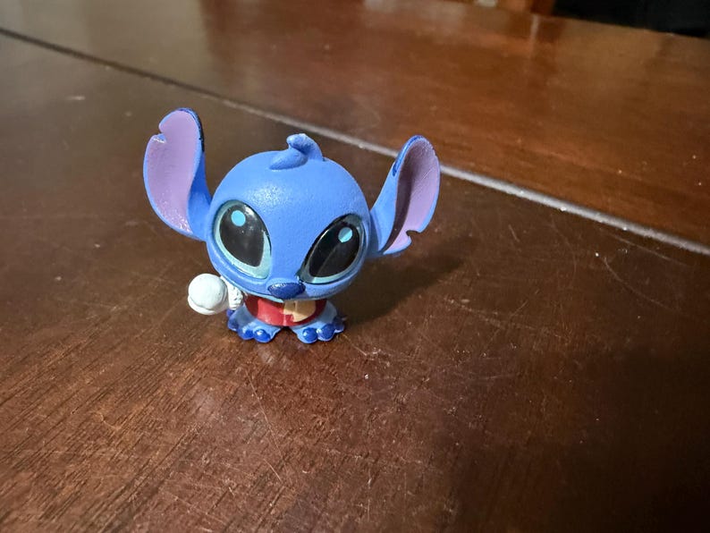 Pu&ograve; includere: Statuetta blu del personaggio Stitch del film d'animazione Lilo & Stitch. La statuetta ha grandi occhi neri, grandi orecchie e una piccola palla bianca in mano. Indossa un colletto rosso.