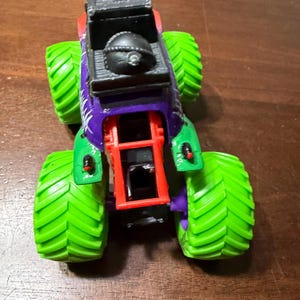 May include: A toy monster truck with a black cab, purple and green body, and bright green tires. The truck has a red chassis and a black detail on top. The tires have a textured pattern.