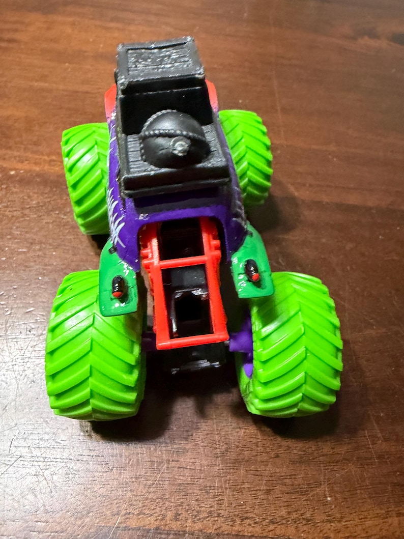 May include: A toy monster truck with a black cab, purple and green body, and bright green tires. The truck has a red chassis and a black detail on top. The tires have a textured pattern.