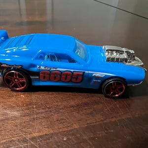 May include: A bright blue toy car with red wheels and a silver engine. The car has the number "6605" and the name "A. Reyna" printed on the side. The car has a spoiler and the words "RIVITED Racing" are printed on the side.