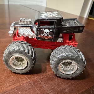 May include: A black and red toy monster truck with large, textured tires. The truck features a skull and crossbones design on the side and top, with silver engine detailing. The chassis is red, and the cab is black.