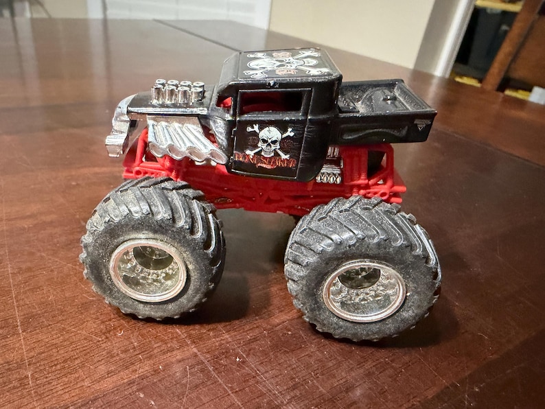 May include: A black and red toy monster truck with large, textured tires. The truck features a skull and crossbones design on the side and top, with silver engine detailing. The chassis is red, and the cab is black.