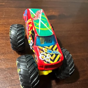 May include: A red and yellow Hot Wheels monster truck toy with black tyres. The truck has a green roof with the Hot Wheels logo and the number 68 on the side. The tyres are large and black with yellow accents.