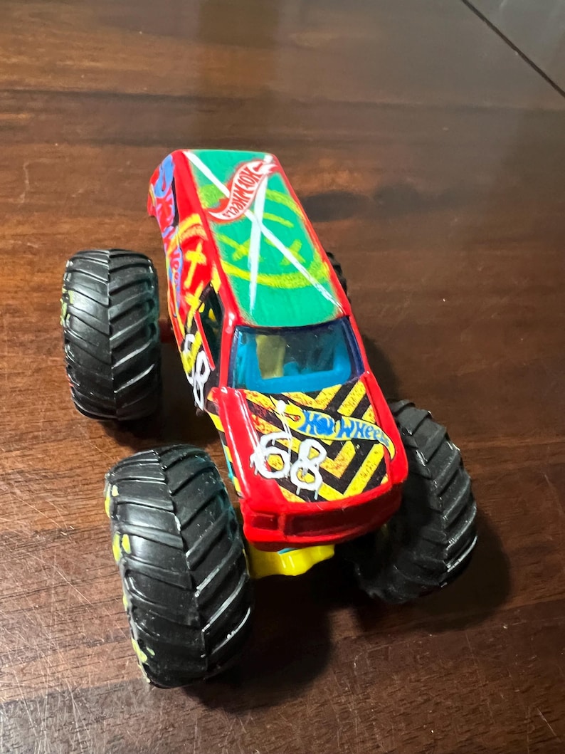 May include: A red and yellow Hot Wheels monster truck toy with black tyres. The truck has a green roof with the Hot Wheels logo and the number 68 on the side. The tyres are large and black with yellow accents.