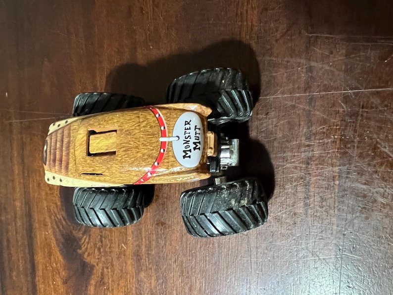 May include: A toy Monster Mutt monster truck with a brown body and black tyres. The truck has a red and white stripe design and the words "Monster Mutt" on the side.