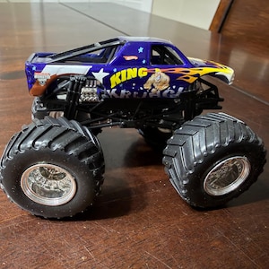 以下が含まれることがあります： 「KING KRUL」の文字が入った、青と黄色のHot Wheelsモンスタートラックのおもちゃ。トラックは、銀色のリムが付いた大きな黒いタイヤと、詳細なエンジンを備えています。