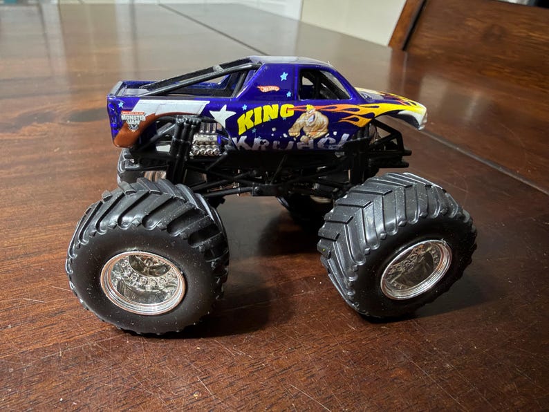 以下が含まれることがあります： 「KING KRUL」の文字が入った、青と黄色のHot Wheelsモンスタートラックのおもちゃ。トラックは、銀色のリムが付いた大きな黒いタイヤと、詳細なエンジンを備えています。