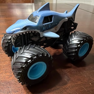 K&ouml;nnte beinhalten: Ein blau-wei&szlig;es Spielzeug-Monstertruck in Form eines Hais, mit dem Wort "MEGALODON" auf der Seite. Der Truck hat gro&szlig;e schwarze Reifen mit blauen Felgen. Das Spielzeug steht auf einer Holzoberfl&auml;che.
