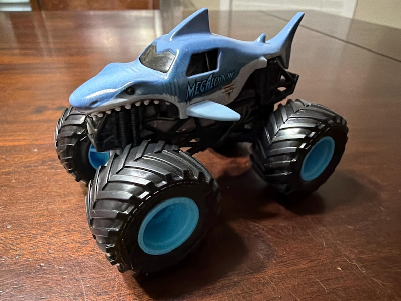 K&ouml;nnte beinhalten: Ein blau-wei&szlig;es Spielzeug-Monstertruck in Form eines Hais, mit dem Wort "MEGALODON" auf der Seite. Der Truck hat gro&szlig;e schwarze Reifen mit blauen Felgen. Das Spielzeug steht auf einer Holzoberfl&auml;che.