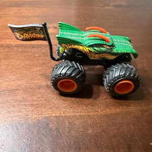 May include: A green and orange monster truck toy with black tires and orange wheel rims. The truck has a dragon design and a flag that reads "DRAGON". The toy is on a brown surface.