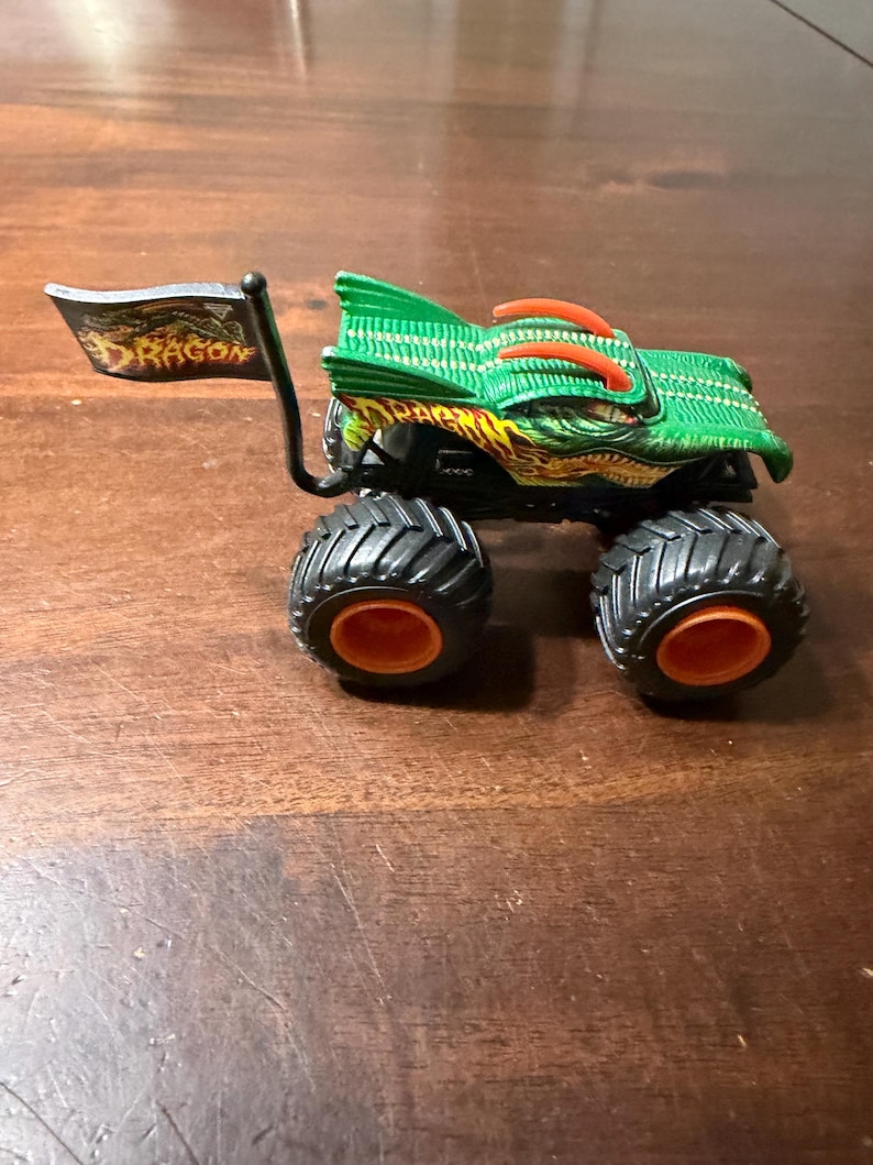 May include: A green and orange monster truck toy with black tires and orange wheel rims. The truck has a dragon design and a flag that reads "DRAGON". The toy is on a brown surface.
