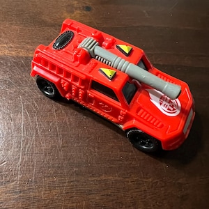 May include: A red toy fire truck with a gray water cannon on top. The truck has black wheels and a white logo on the side.