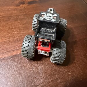 May include: A black and red toy monster truck with large, textured tires. The truck bed features a skull and crossbones design and the words "Bone Shaker". The toy is designed for play and collecting.