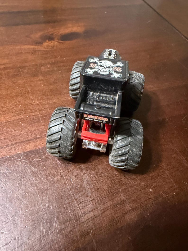May include: A black and red toy monster truck with large, textured tires. The truck bed features a skull and crossbones design and the words "Bone Shaker". The toy is designed for play and collecting.