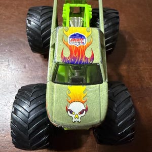 Puede incluir: Un cami&oacute;n monstruo de juguete verde con neum&aacute;ticos negros grandes. El cami&oacute;n presenta un gr&aacute;fico de calavera con llamas y el logotipo "LUCAS OIL PRODUCTS INC.". El cami&oacute;n tiene una jaula antivuelco verde y un parabrisas negro.