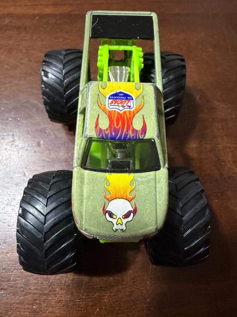 Puede incluir: Un cami&oacute;n monstruo de juguete verde con neum&aacute;ticos negros grandes. El cami&oacute;n presenta un gr&aacute;fico de calavera con llamas y el logotipo "LUCAS OIL PRODUCTS INC.". El cami&oacute;n tiene una jaula antivuelco verde y un parabrisas negro.