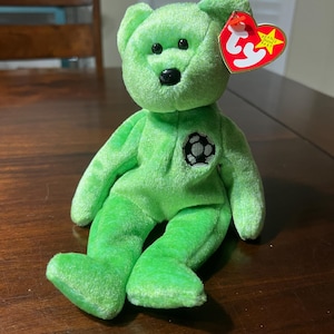 Puede incluir: Un oso de peluche verde con un emblema de balón de fútbol en el pecho. El oso tiene ojos negros y una nariz negra. Una etiqueta roja en forma de corazón está unida a su oreja.
