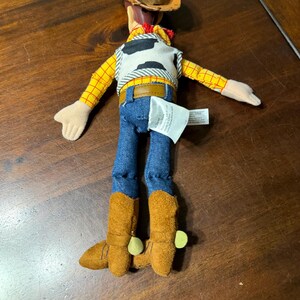 May include: A plush toy of a cowboy character, featuring a brown hat, yellow plaid shirt, blue jeans, and brown boots. The vest is white with black spots. The toy has a red neckerchief and a brown belt.