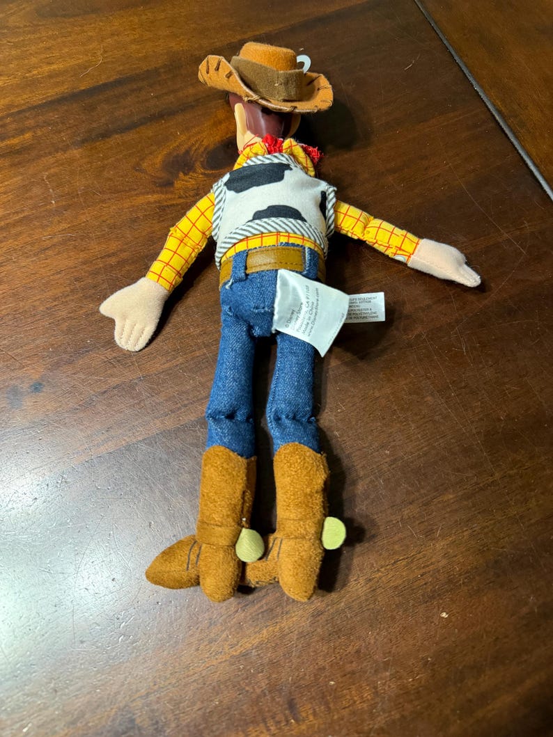 May include: A plush toy of a cowboy character, featuring a brown hat, yellow plaid shirt, blue jeans, and brown boots. The vest is white with black spots. The toy has a red neckerchief and a brown belt.